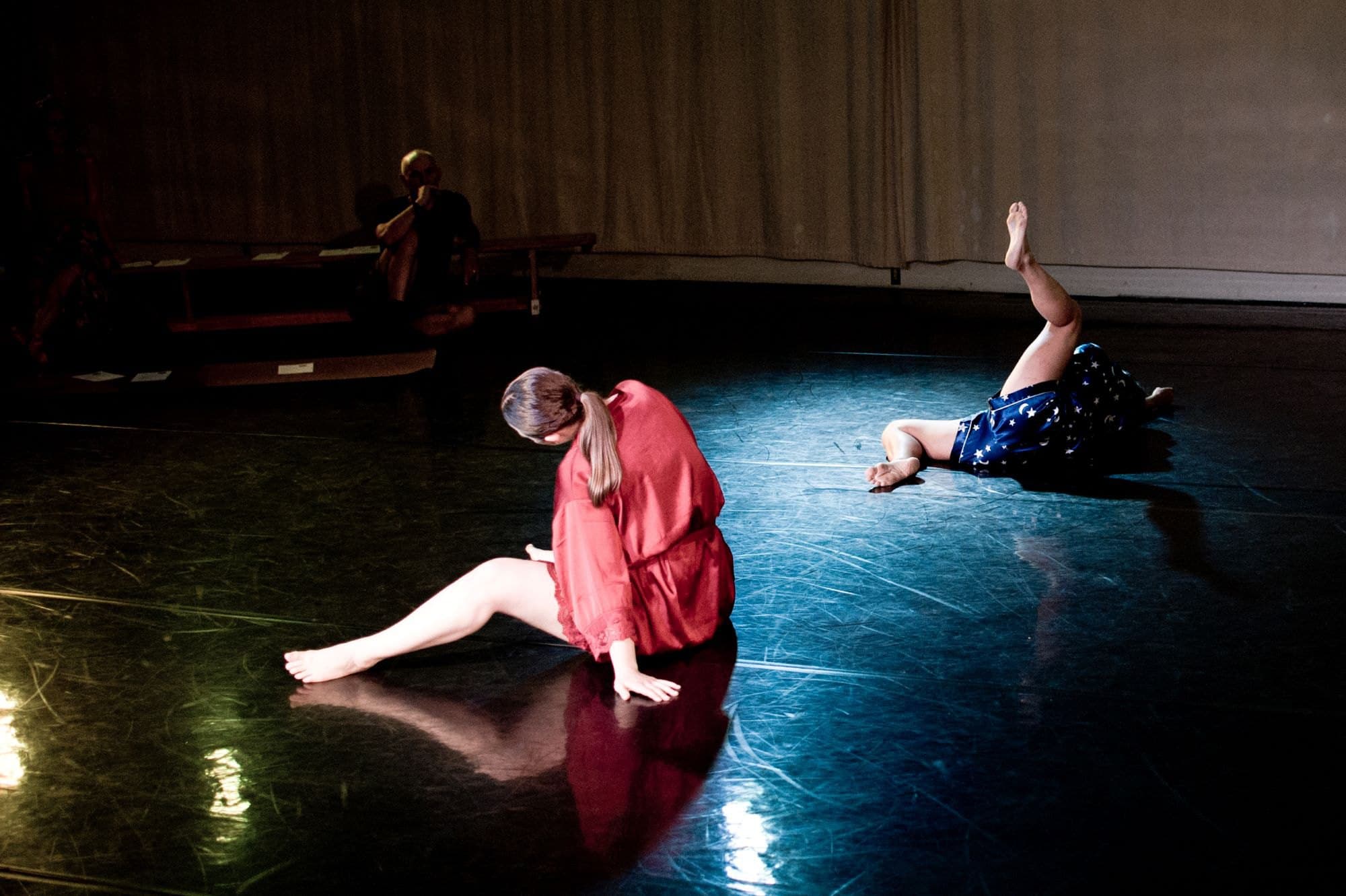 Two dancers in pyjamas on a dark stage, one in red sitting, one in blue lying back