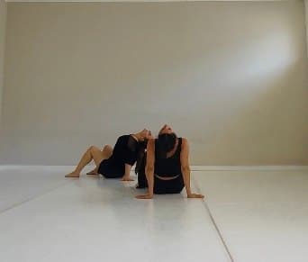 Two dancers in black leaning back to back on a white floor